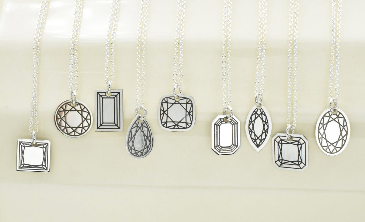 A collection of silver geometric pendant necklaces with clear faceted gemstones, displayed on a cream-colored background.